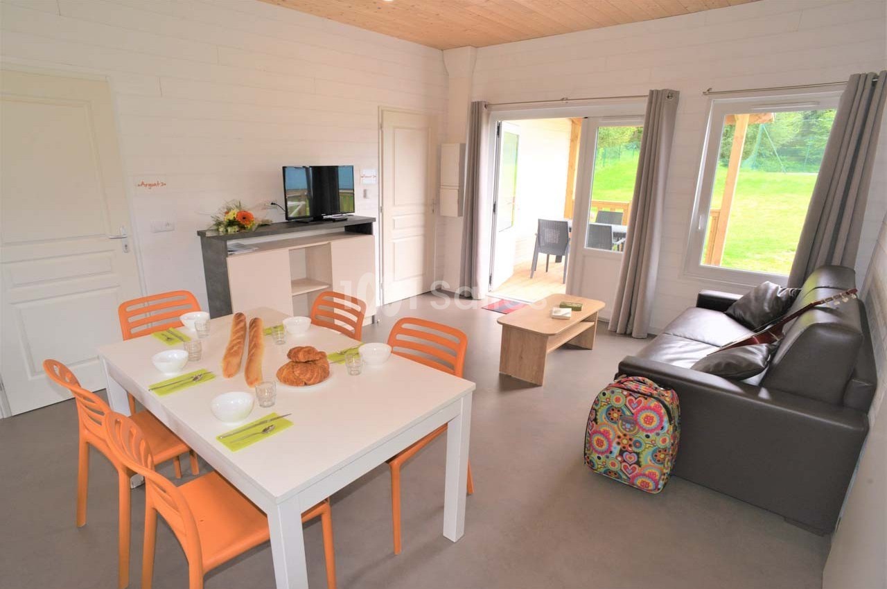 Salle de séjour lumineuse avec table à manger, canapé, télévision, et vue sur un jardin à travers une baie vitrée.