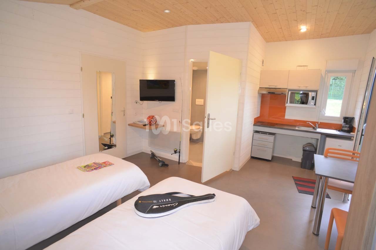Intérieur d'un chalet avec deux lits simples, une kitchenette équipée, une table et une télévision fixée au mur.