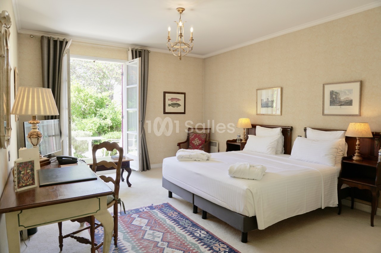 Chambre lumineuse avec lit double, bureau, fauteuil, tapis coloré et fenêtre ouverte sur un jardin verdoyant.