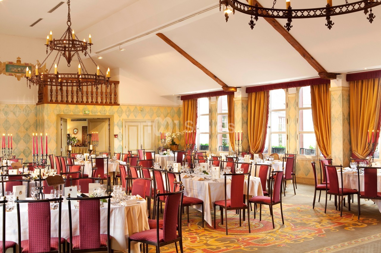 Salle de réception élégante avec tables dressées, chaises rouges, lustres en fer forgé et grandes fenêtres avec rideaux…