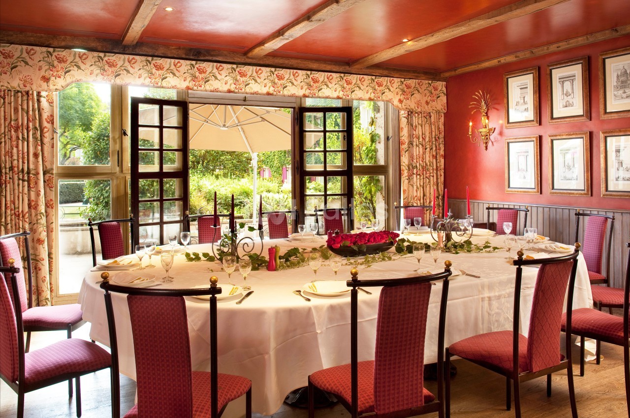 Salle à manger élégante avec une grande table dressée, entourée de chaises rouges, et vue sur un jardin par des fenêtres…