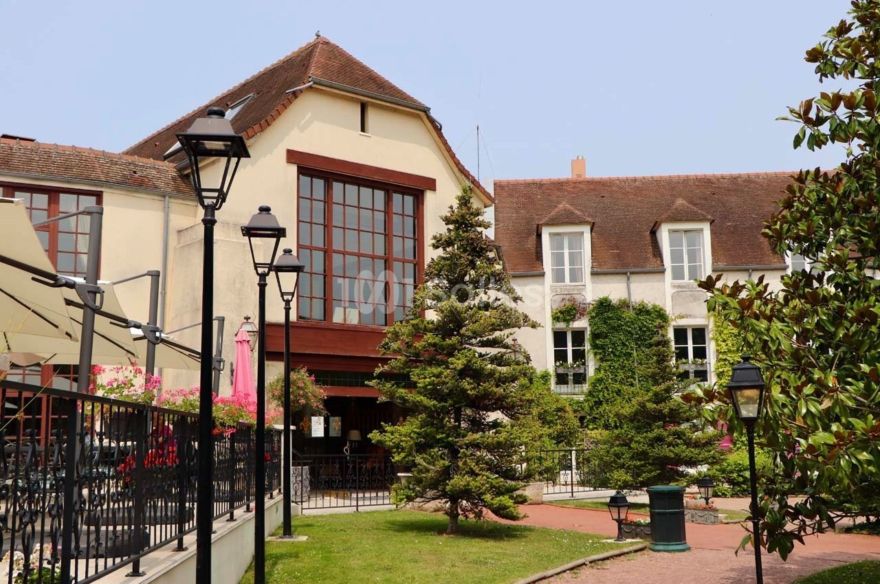 Façade d'un bâtiment avec grandes fenêtres, terrasse ombragée, lampadaires et jardin arboré au premier plan.