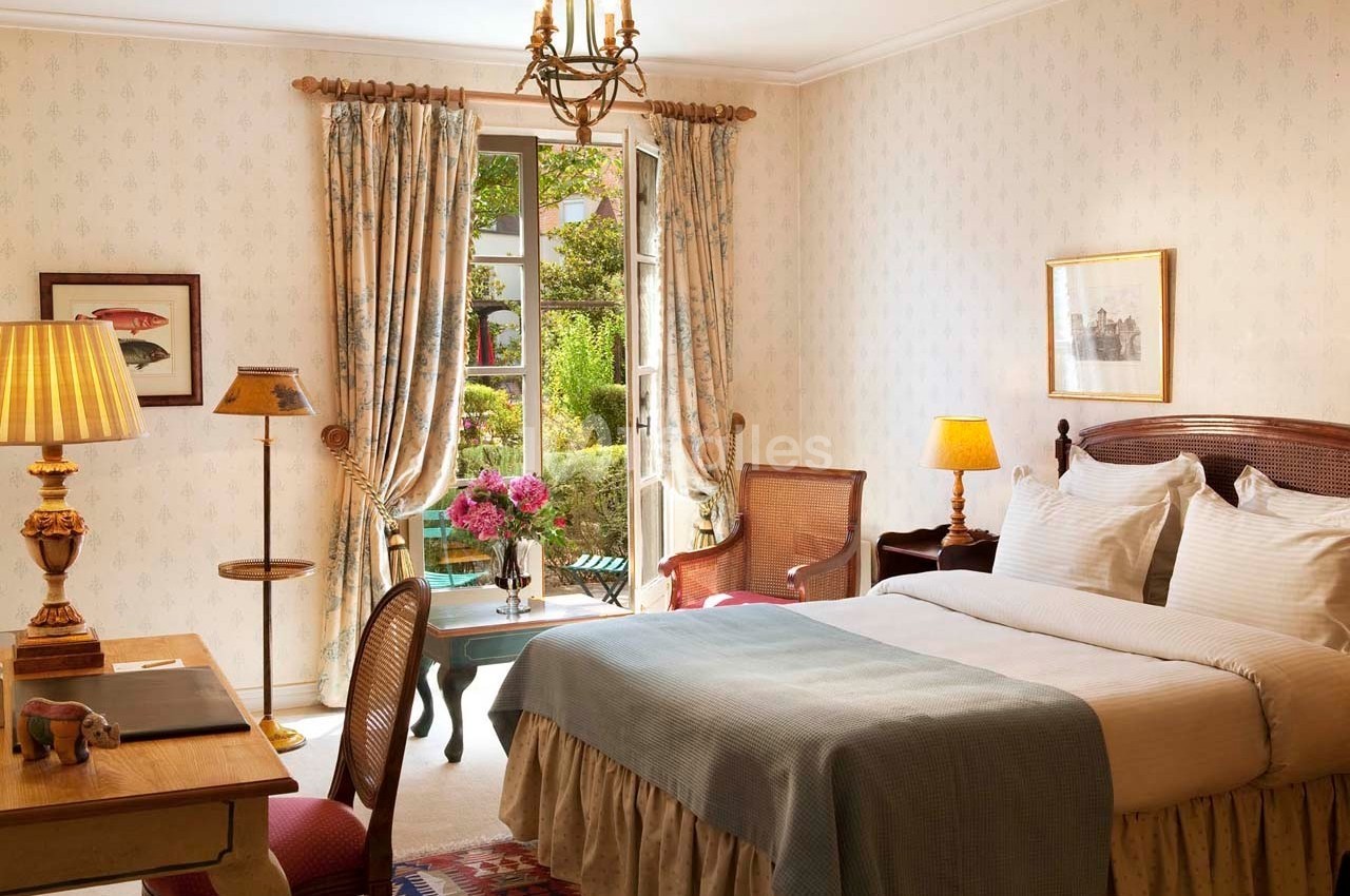 Chambre lumineuse avec lit double, bureau en bois, fauteuils et vue sur un jardin à travers une porte-fenêtre ouverte.