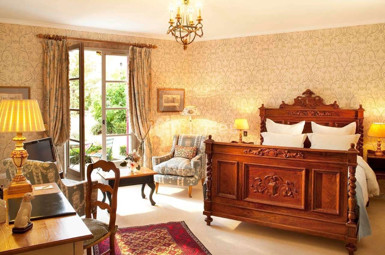 Chambre lumineuse avec lit en bois sculpté, bureau, fauteuil, tapis rouge et vue sur un jardin par une porte-fenêtre.