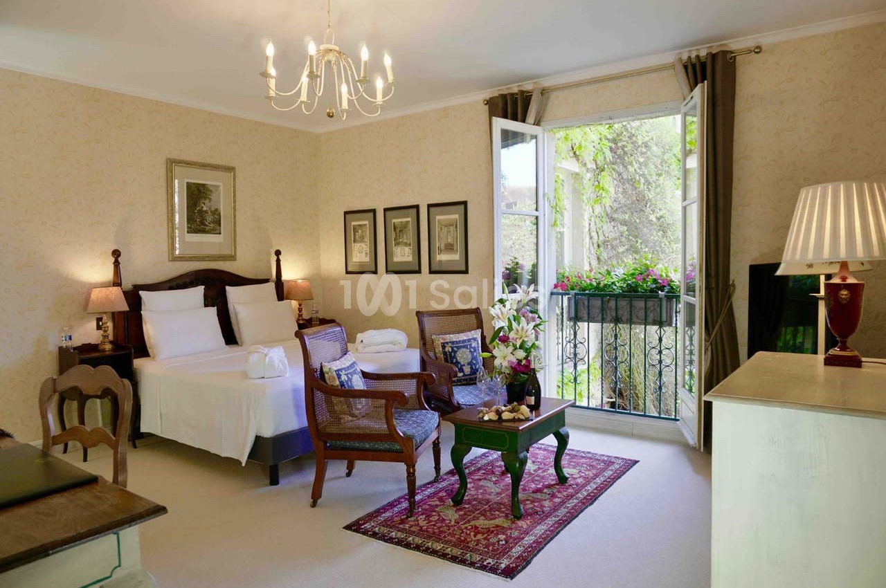 Chambre lumineuse avec lit double, coin salon, balcon ouvert sur un jardin et décoration classique.