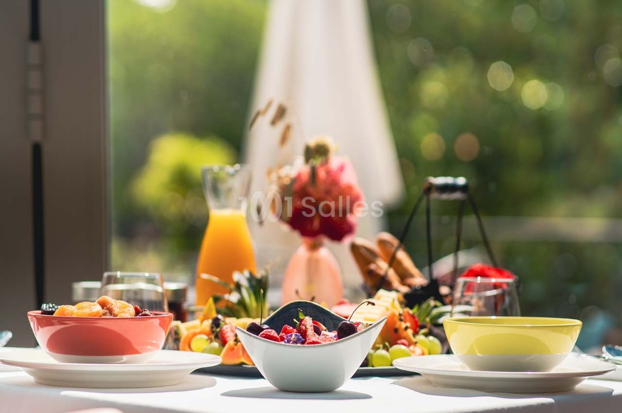 Table dressée avec des bols de fruits, un pichet de jus d'orange et des décorations florales en arrière-plan lumineux.