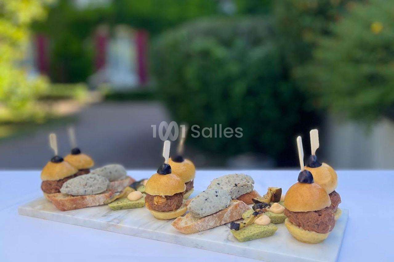 Plateau de mini-burgers et amuse-bouches variés présentés sur une table en extérieur, avec un jardin en arrière-plan.