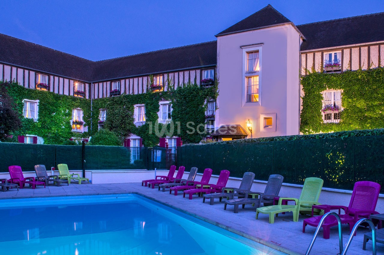 Piscine extérieure entourée de transats colorés devant un bâtiment à colombages éclairé en soirée.
