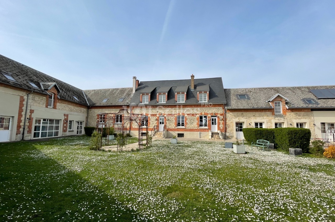 Grande maison en pierre avec toit en ardoise, entourée d'une cour herbeuse parsemée de petites fleurs blanches.