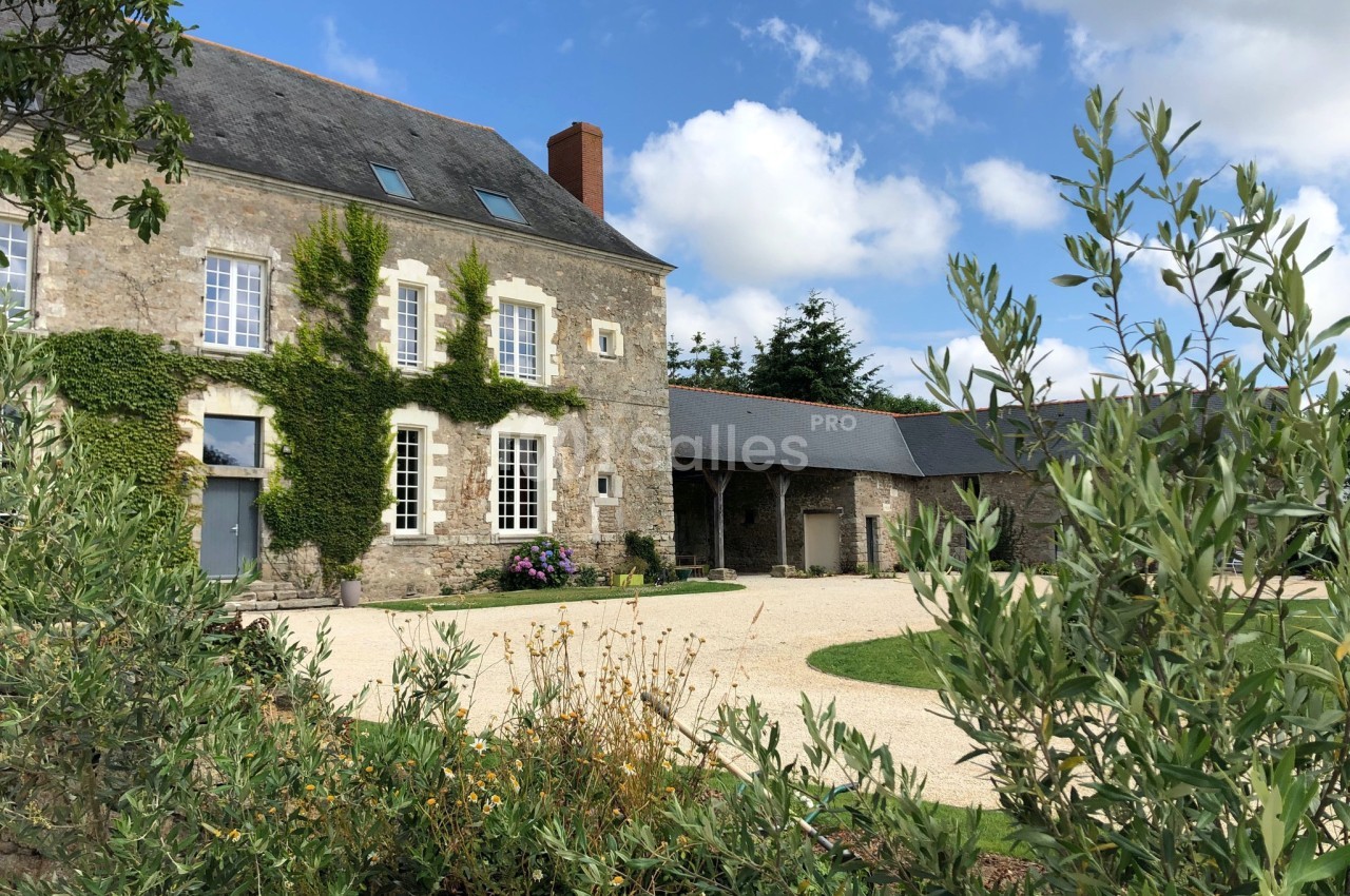 Maison en pierre avec façade végétalisée, entourée d'une cour gravillonnée et de végétation sous un ciel partiellement…