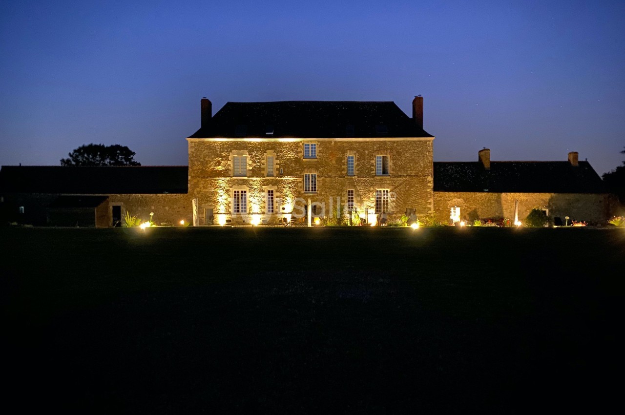 Façade d'une grande maison en pierre éclairée la nuit, entourée d'un jardin sombre et d'annexes.