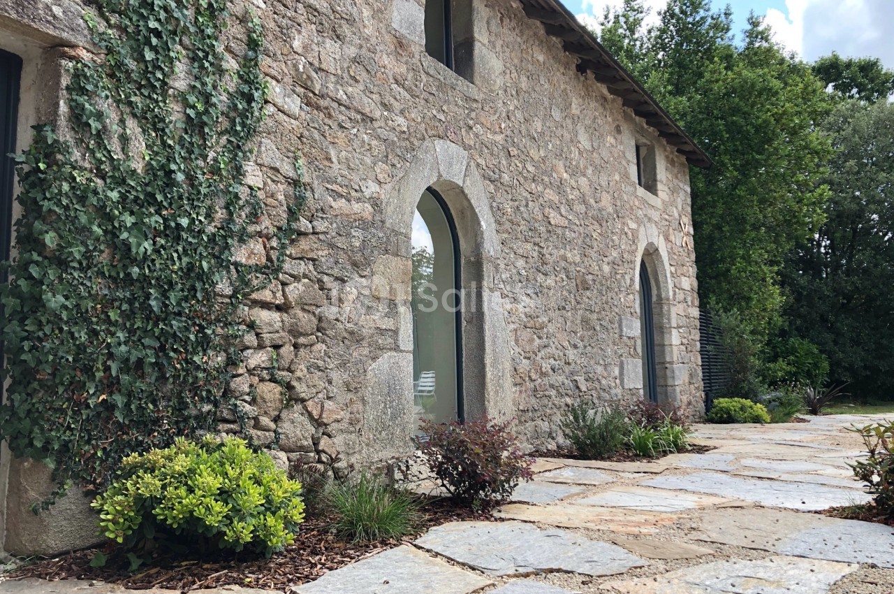 Façade en pierre d'une maison ancienne avec des arches, entourée de végétation et d'un sol en dalles.