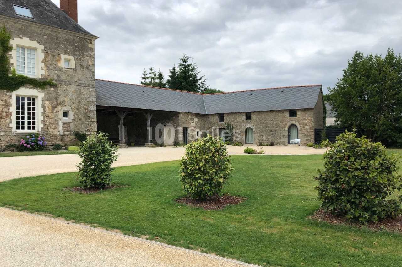 Bâtisse en pierre avec toit en ardoise, entourée d'une pelouse et de buissons, sous un ciel nuageux.