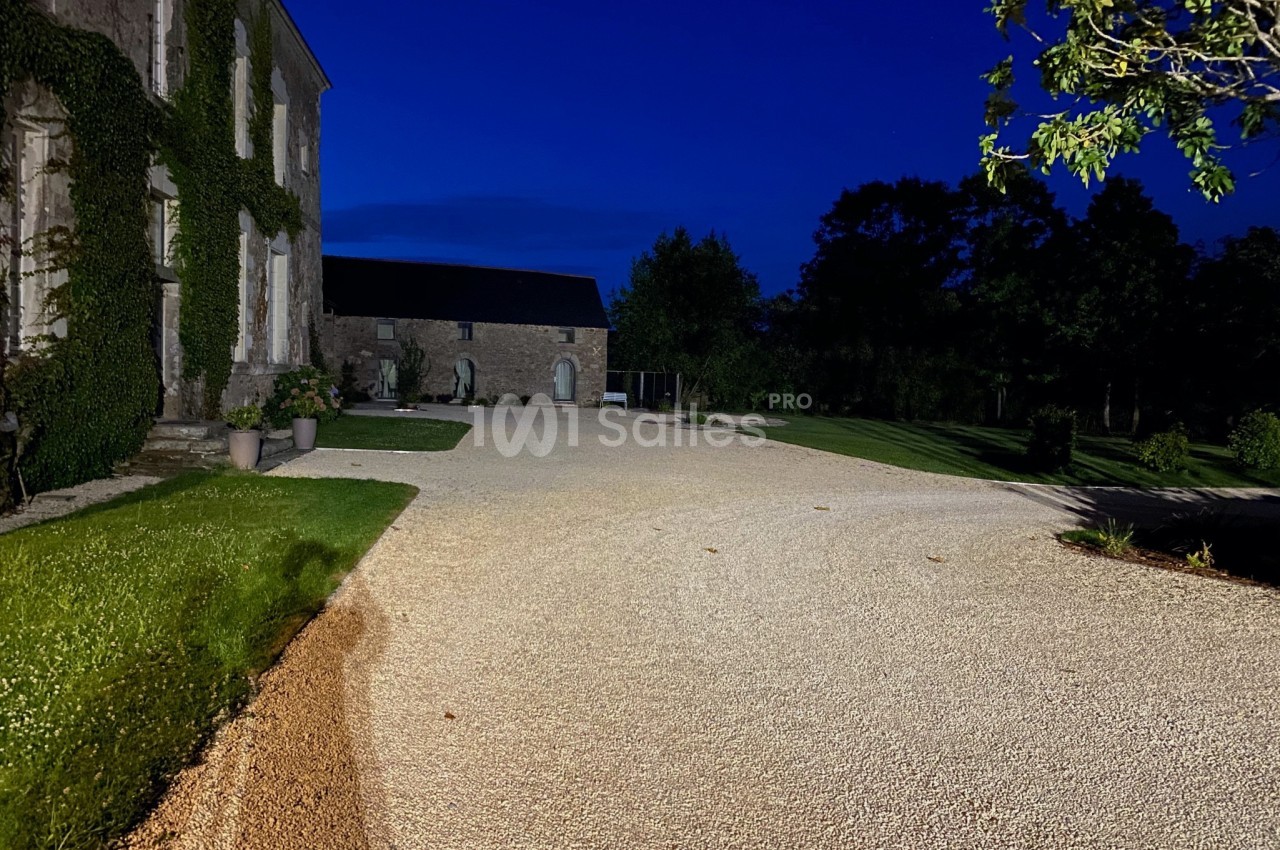 Allée en gravier éclairée la nuit, bordée de pelouses et d'un bâtiment en pierre recouvert de végétation.