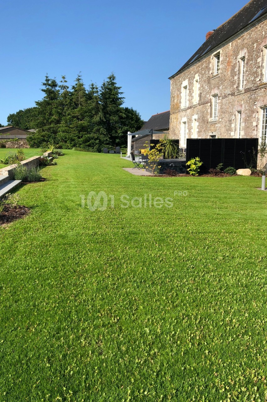 Pelouse bien entretenue devant une maison en pierre avec terrasse et arbres en arrière-plan.