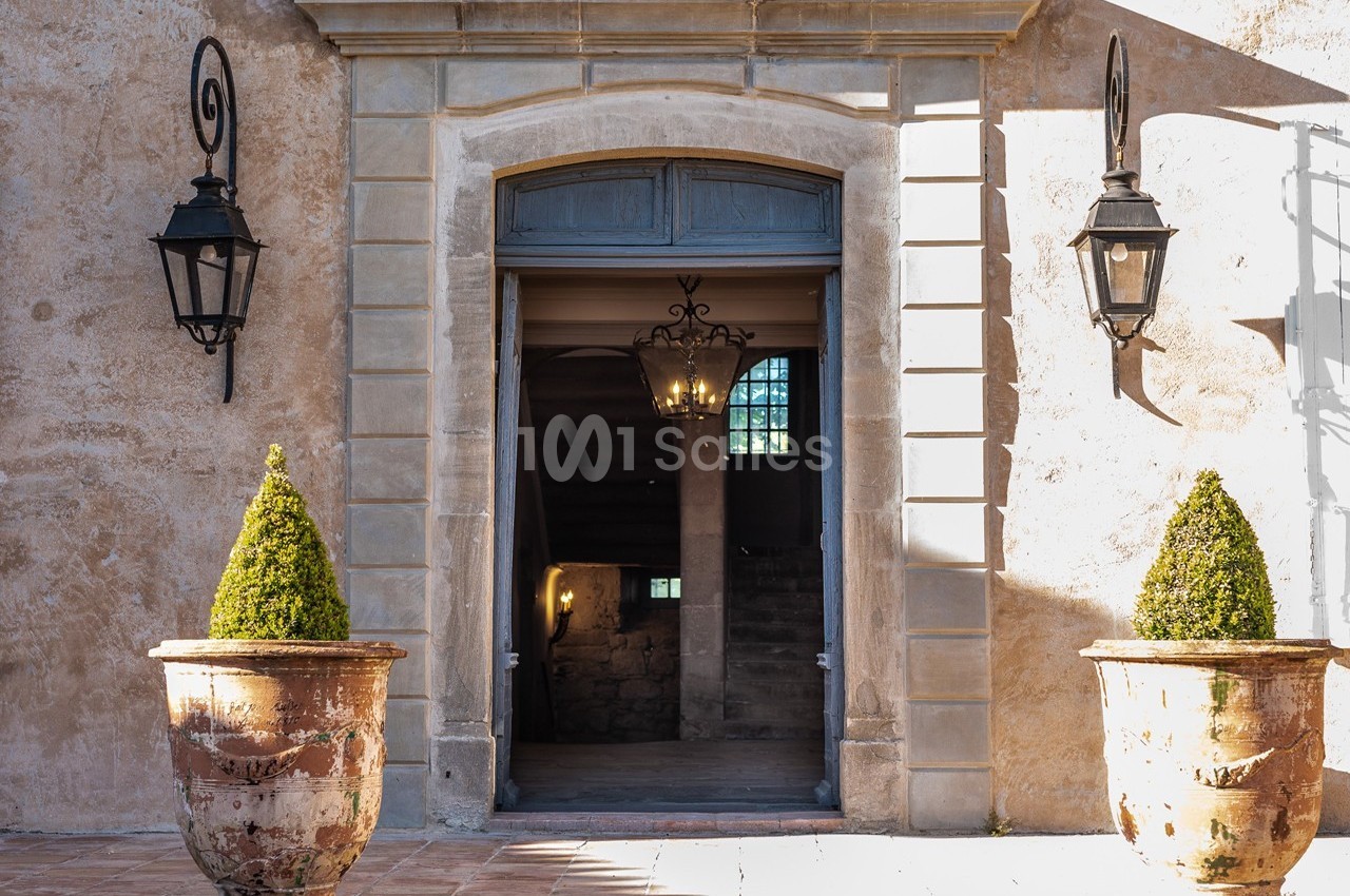 Entrée d'un bâtiment ancien avec porte ouverte, deux lanternes murales et deux pots en terre cuite contenant des arbustes…