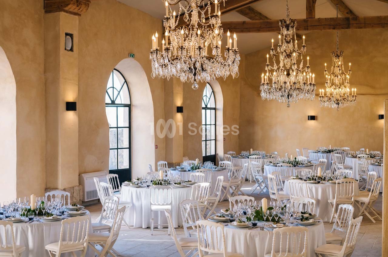 Salle de réception élégante avec murs beige, grandes fenêtres cintrées, lustres en cristal et tables rondes dressées.