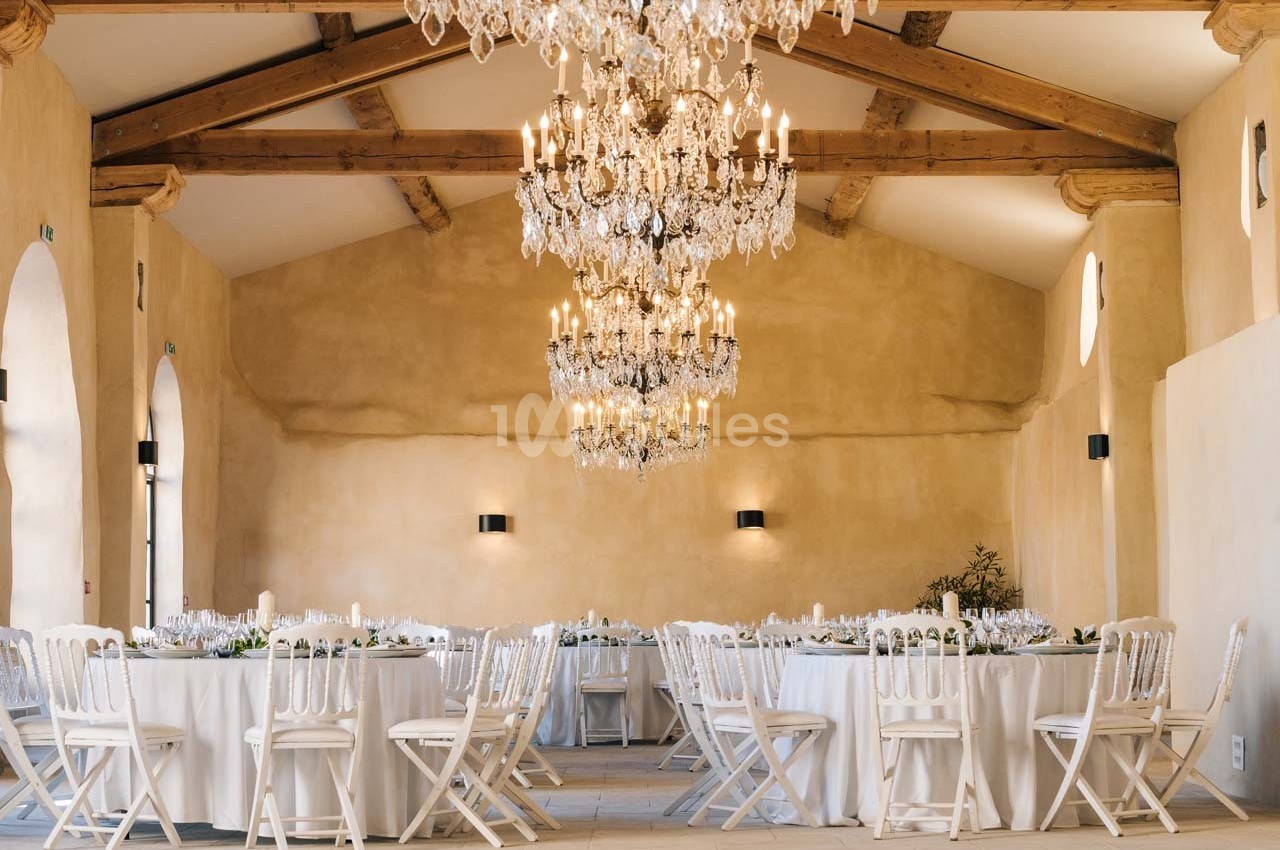 Salle de réception lumineuse avec tables rondes dressées, chaises blanches et lustres en cristal suspendus.