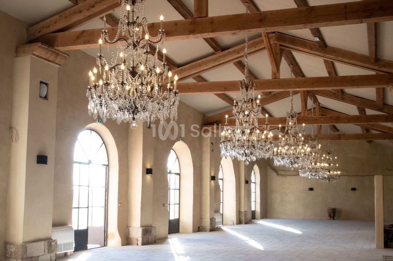 Salle spacieuse avec poutres apparentes, lustres en cristal suspendus et grandes fenêtres cintrées laissant entrer la…