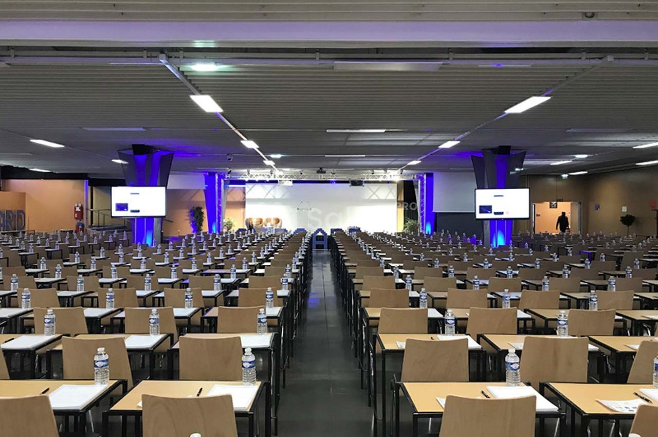 Salle de conférence avec de nombreuses rangées de tables et chaises, équipée d'écrans et de bouteilles d'eau sur chaque…