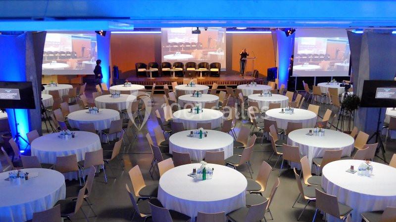 Salle de conférence avec des tables rondes dressées, des chaises disposées et une scène équipée d'un podium.