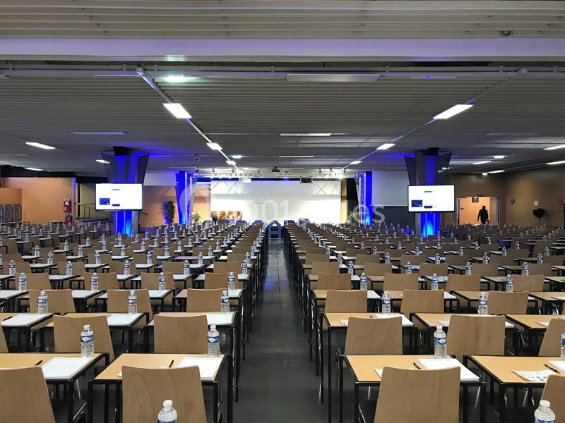 Salle de conférence avec rangées de tables et chaises, écrans allumés et bouteilles d'eau disposées sur les tables.
