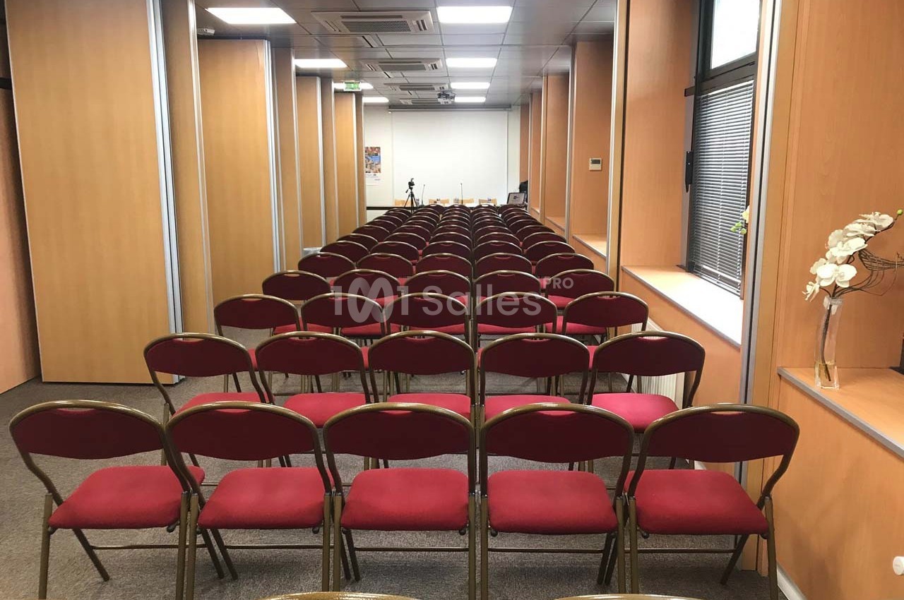 Salle de conférence avec rangées de chaises rouges alignées face à un écran blanc.