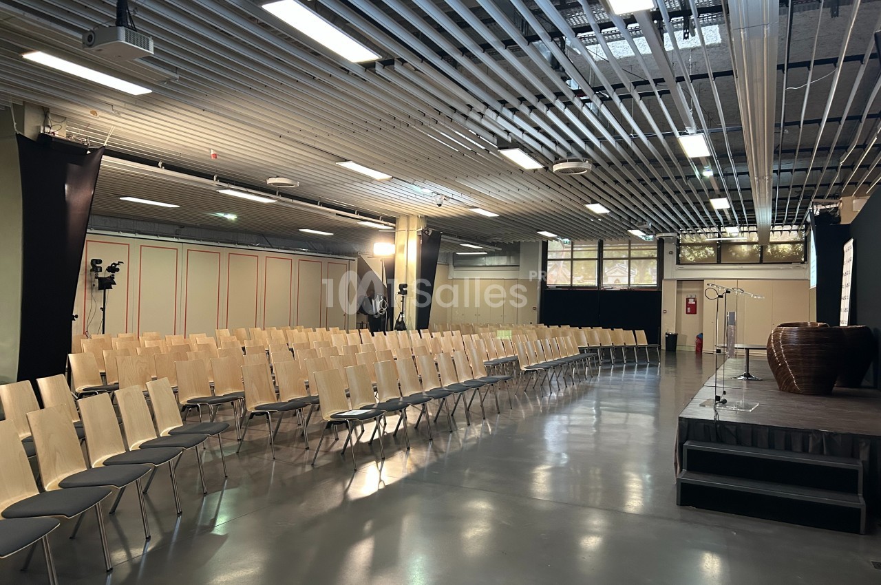 Salle de conférence vide avec des rangées de chaises en bois, une estrade et des équipements audiovisuels.
