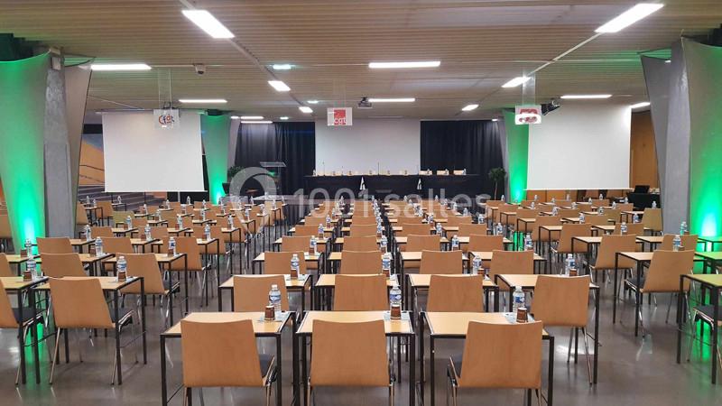Salle de conférence aménagée avec des rangées de tables et chaises, équipée d'un podium et d'écrans de projection.