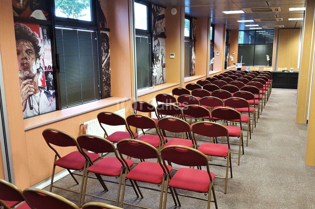 Salle de conférence avec des rangées de chaises rouges alignées devant des fenêtres et des murs décorés.