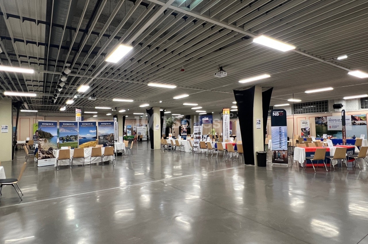 Salle d'exposition avec des stands alignés sous un plafond industriel, éclairée par des lumières artificielles.