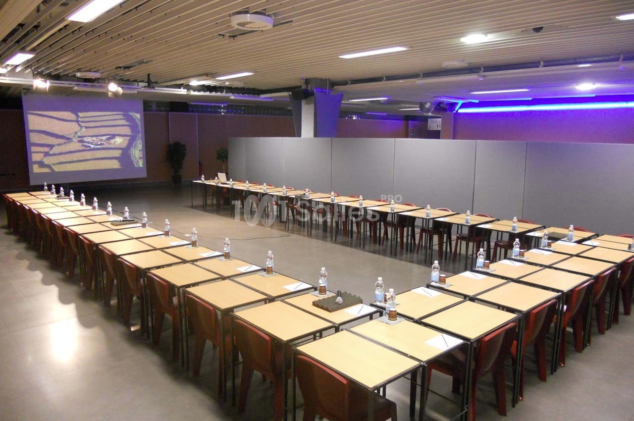 Salle de conférence avec des tables disposées en U, équipées de bouteilles d'eau et de documents, éclairage moderne.