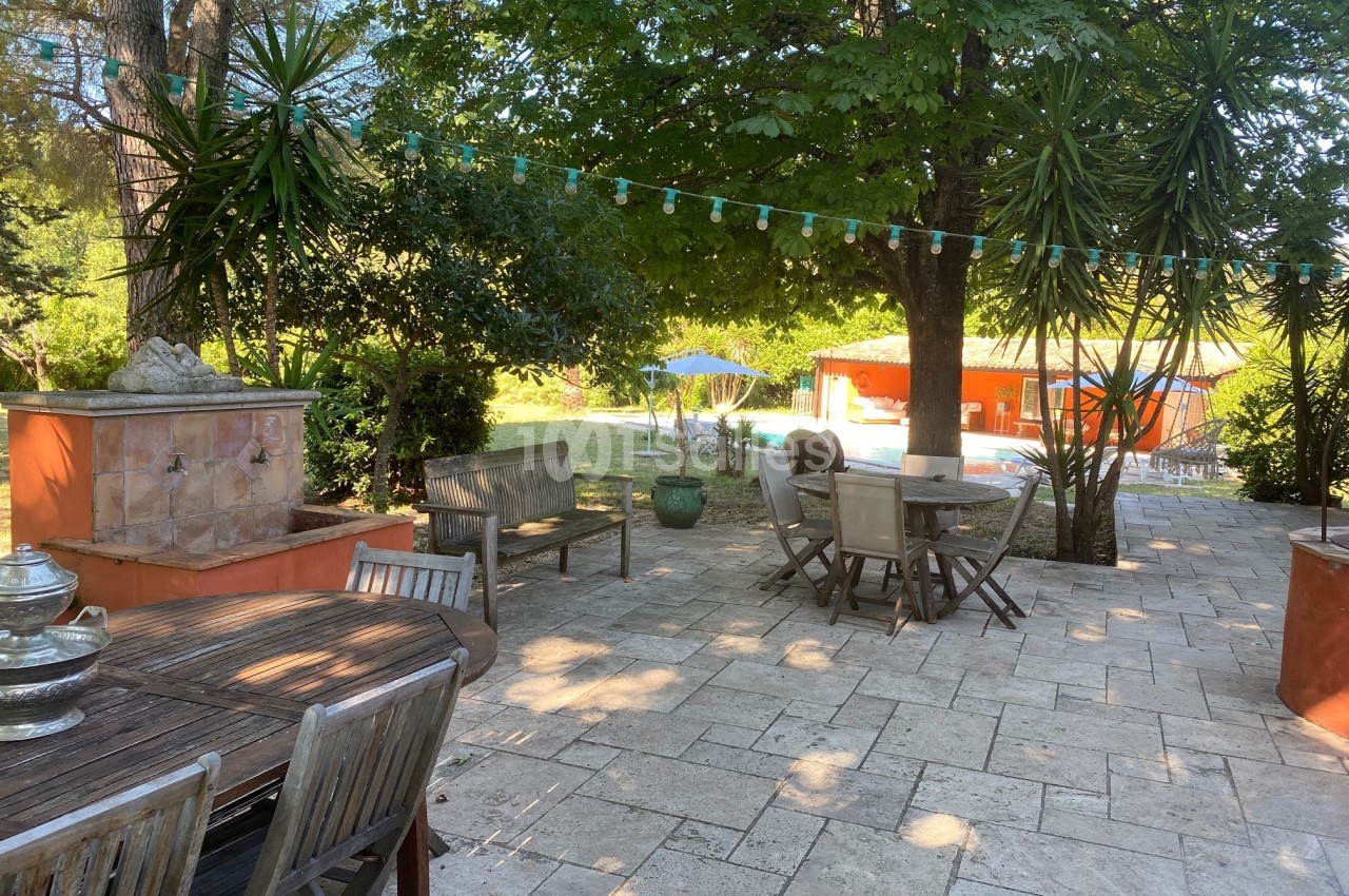 Terrasse en pierre ombragée avec mobilier de jardin, guirlande lumineuse et vue sur une piscine entourée de verdure.