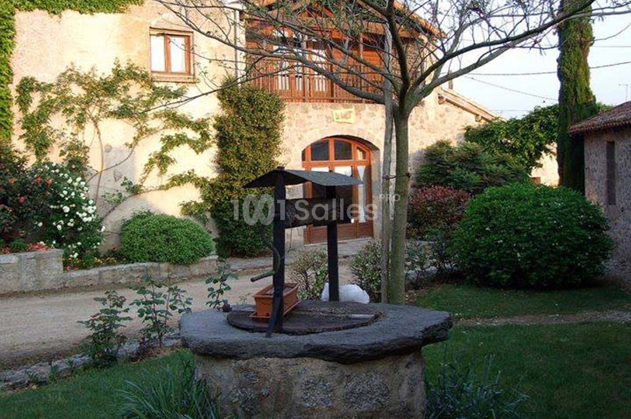 Vieille maison en pierre avec un puits en premier plan, entourée de verdure et de fleurs.