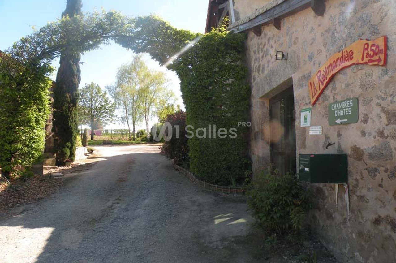 Entrée d'une maison en pierre avec une arche végétale, un panneau indiquant des chambres d'hôtes et un chemin gravillonné.