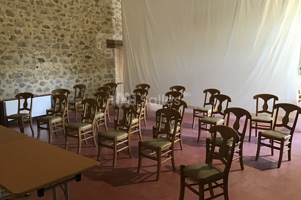 Salle avec des chaises en bois disposées en rangées devant un mur en pierre et un grand drap blanc tendu.