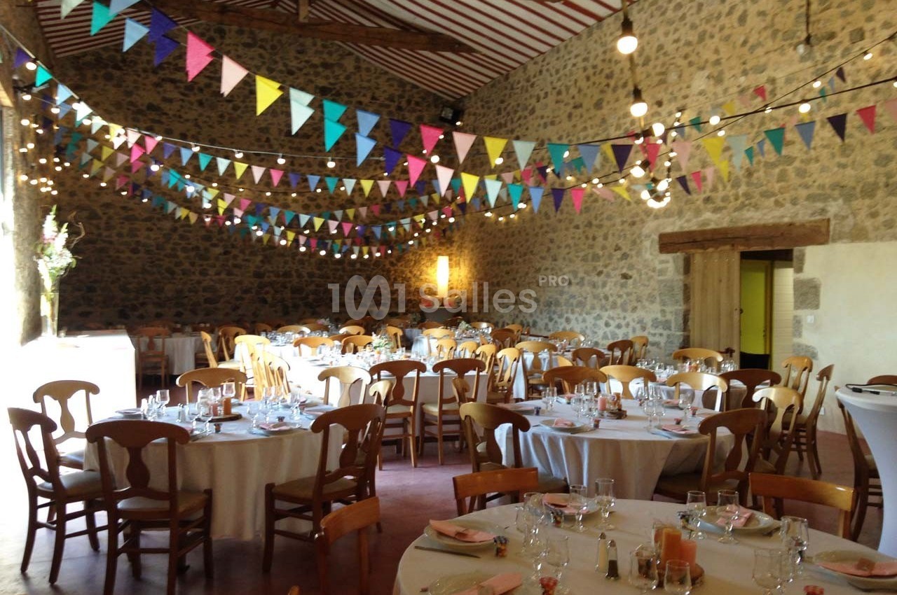 Salle de réception décorée de guirlandes colorées, avec des tables rondes dressées pour un événement festif.