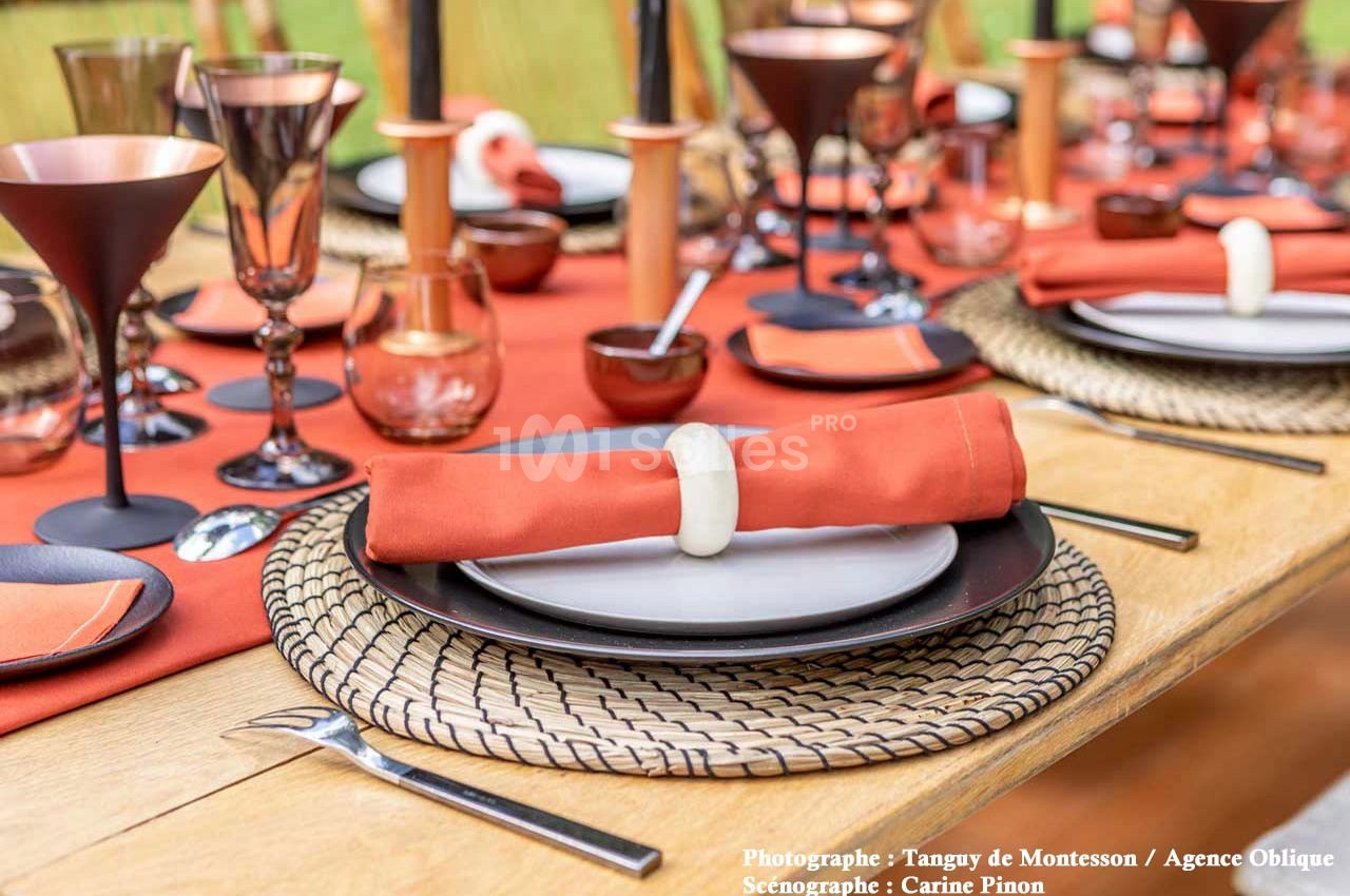 Table dressée avec des assiettes noires, serviettes orange nouées, verres en cuivre et décoration élégante sur nappe rouge.