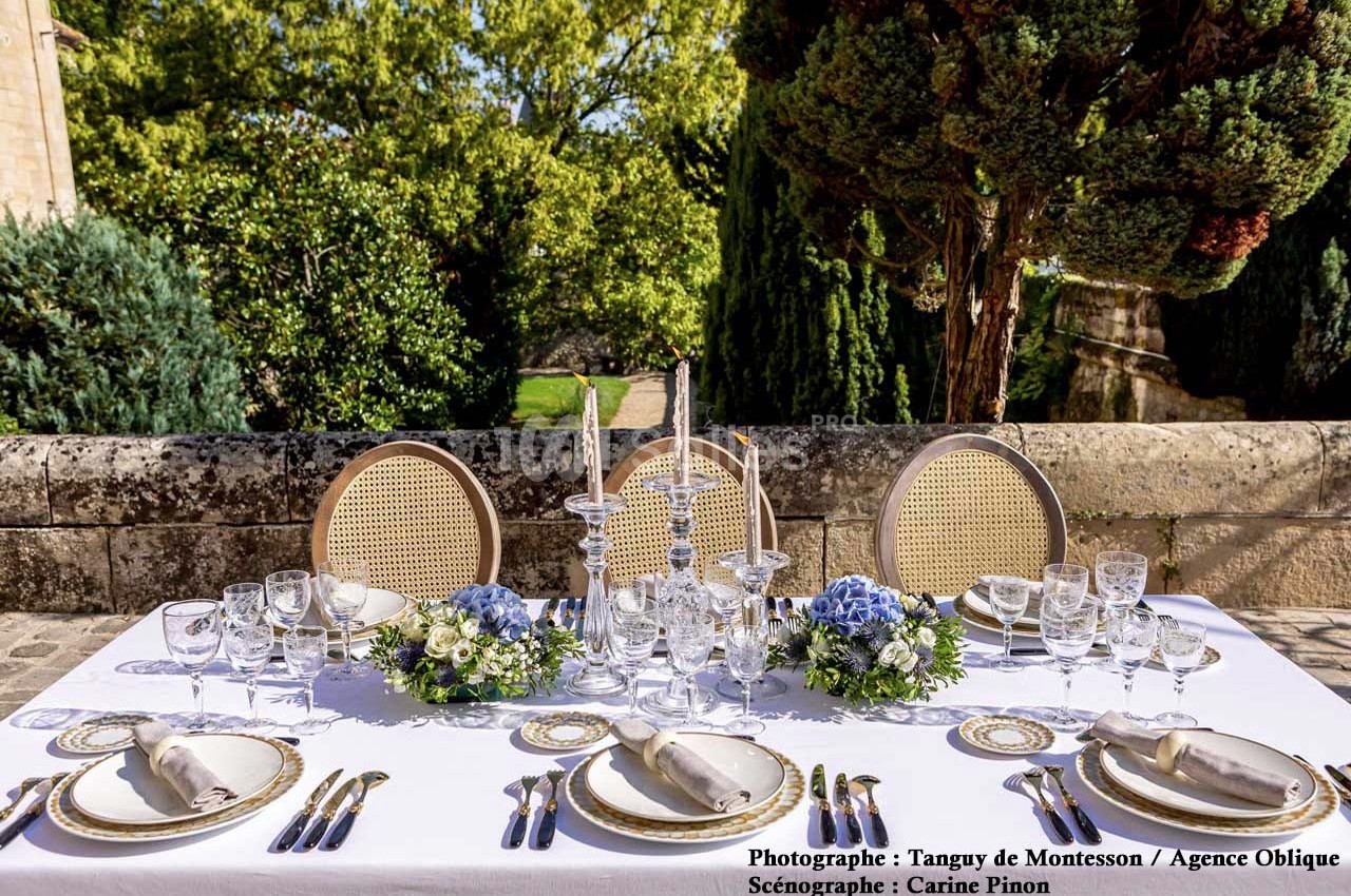 Table élégamment dressée en extérieur avec vaisselle raffinée, compositions florales et vue sur un jardin arboré.