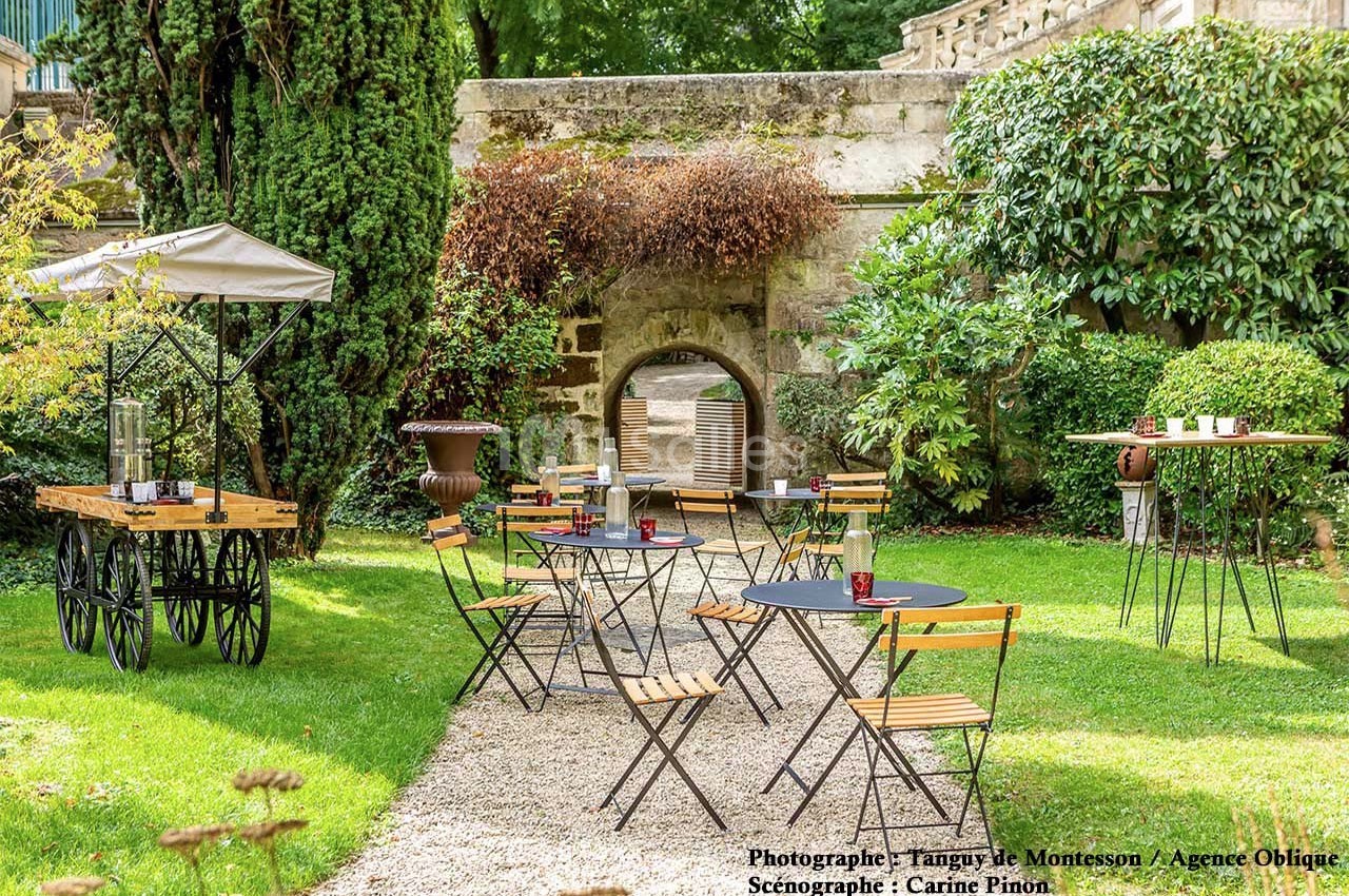 Jardin aménagé avec tables et chaises en métal et bois, entouré de verdure et d'une arche en pierre au fond.