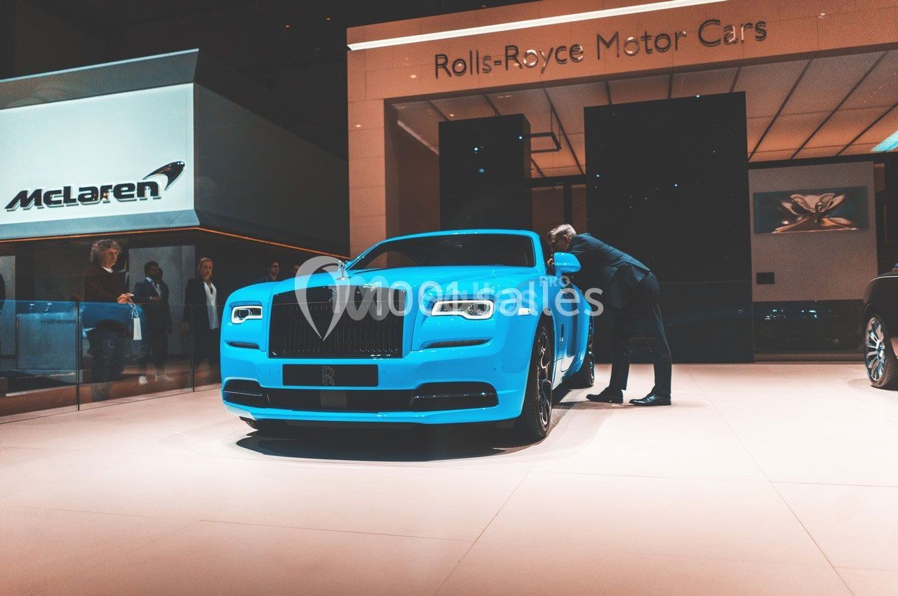 Une voiture de luxe bleue exposée dans un showroom, un homme examine l'intérieur par la portière ouverte.