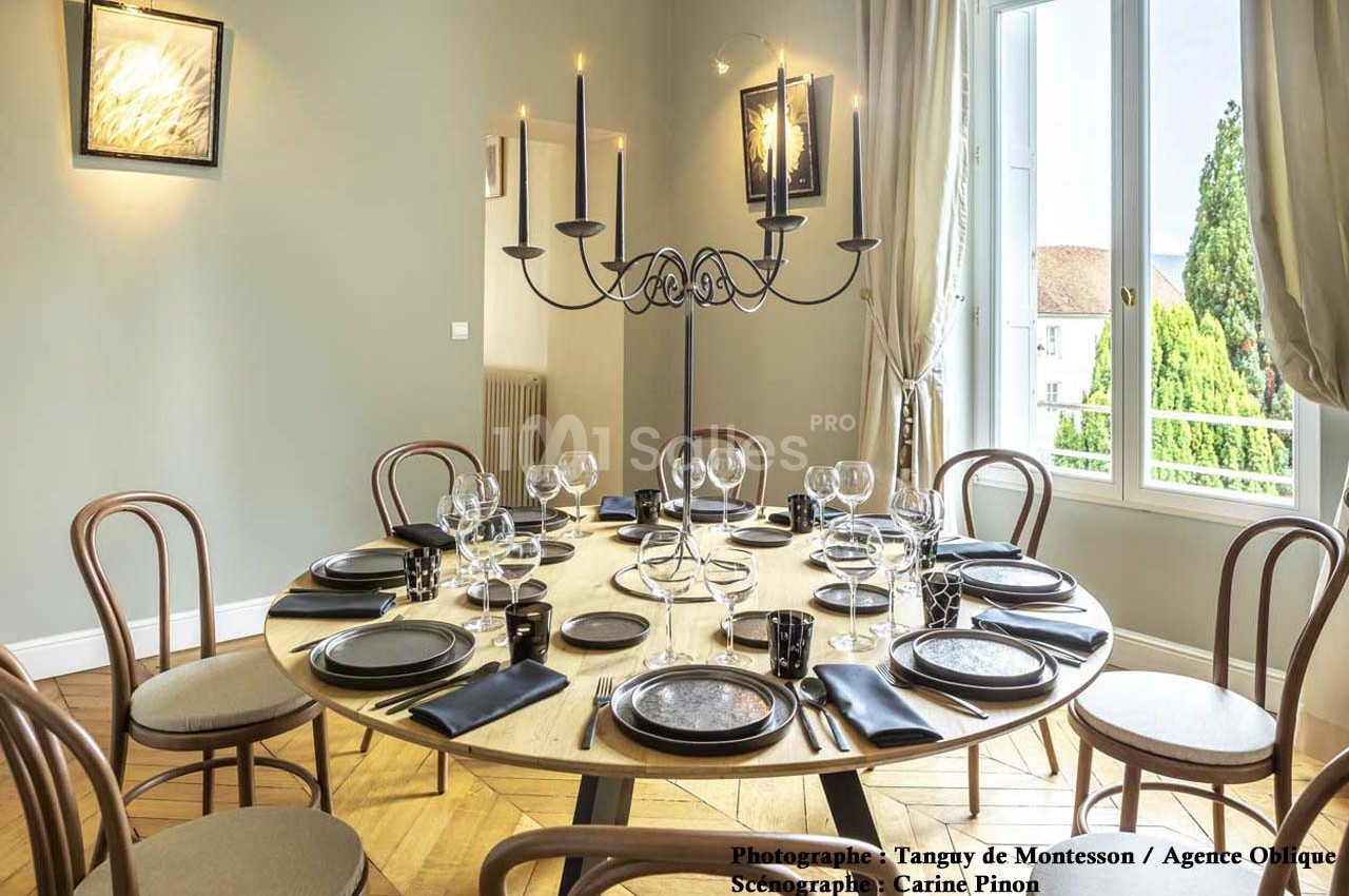 Table ronde élégamment dressée avec vaisselle noire, chandelier central et vue sur un paysage extérieur par une grande…