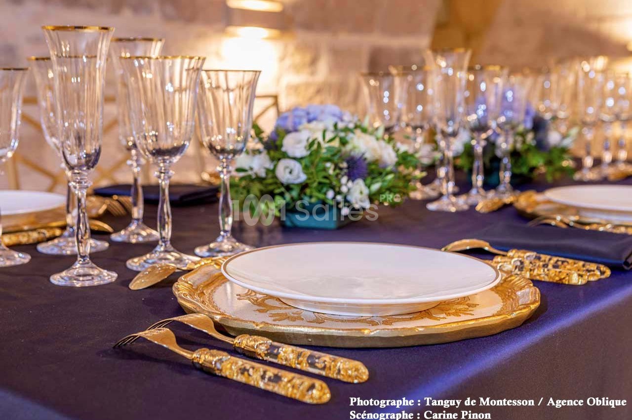 Table élégamment dressée avec vaisselle dorée, verres en cristal et centre de table floral sur une nappe sombre.