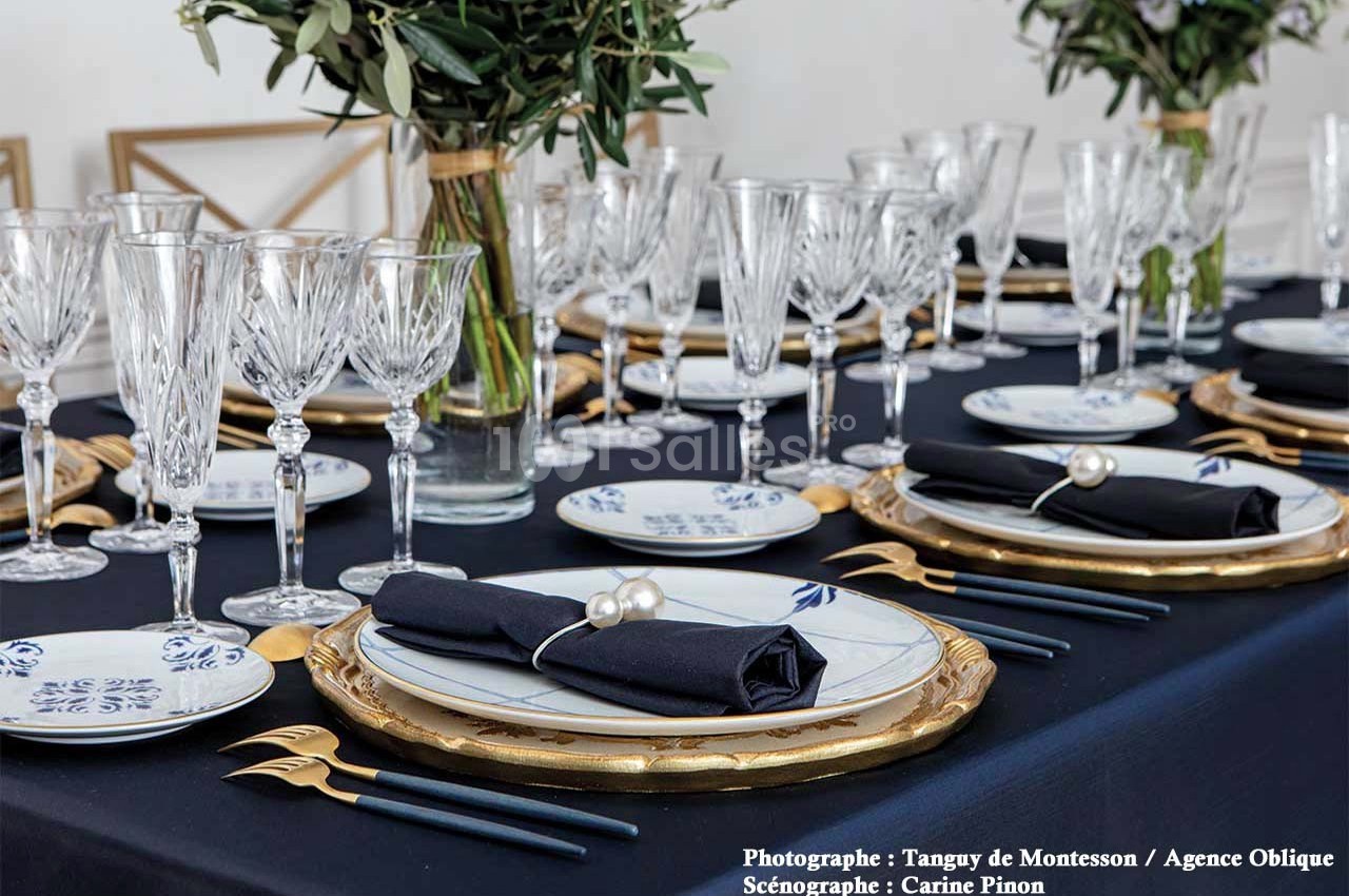 Table élégamment dressée avec nappes noires, vaisselle blanche et dorée, couverts dorés et verres en cristal.