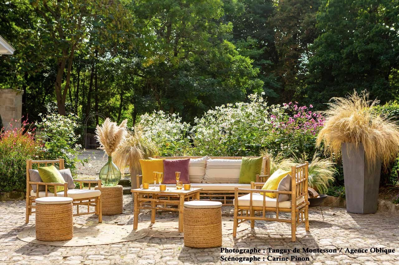 Salon de jardin en rotin avec coussins colorés, entouré de végétation et de fleurs, sur une terrasse pavée.