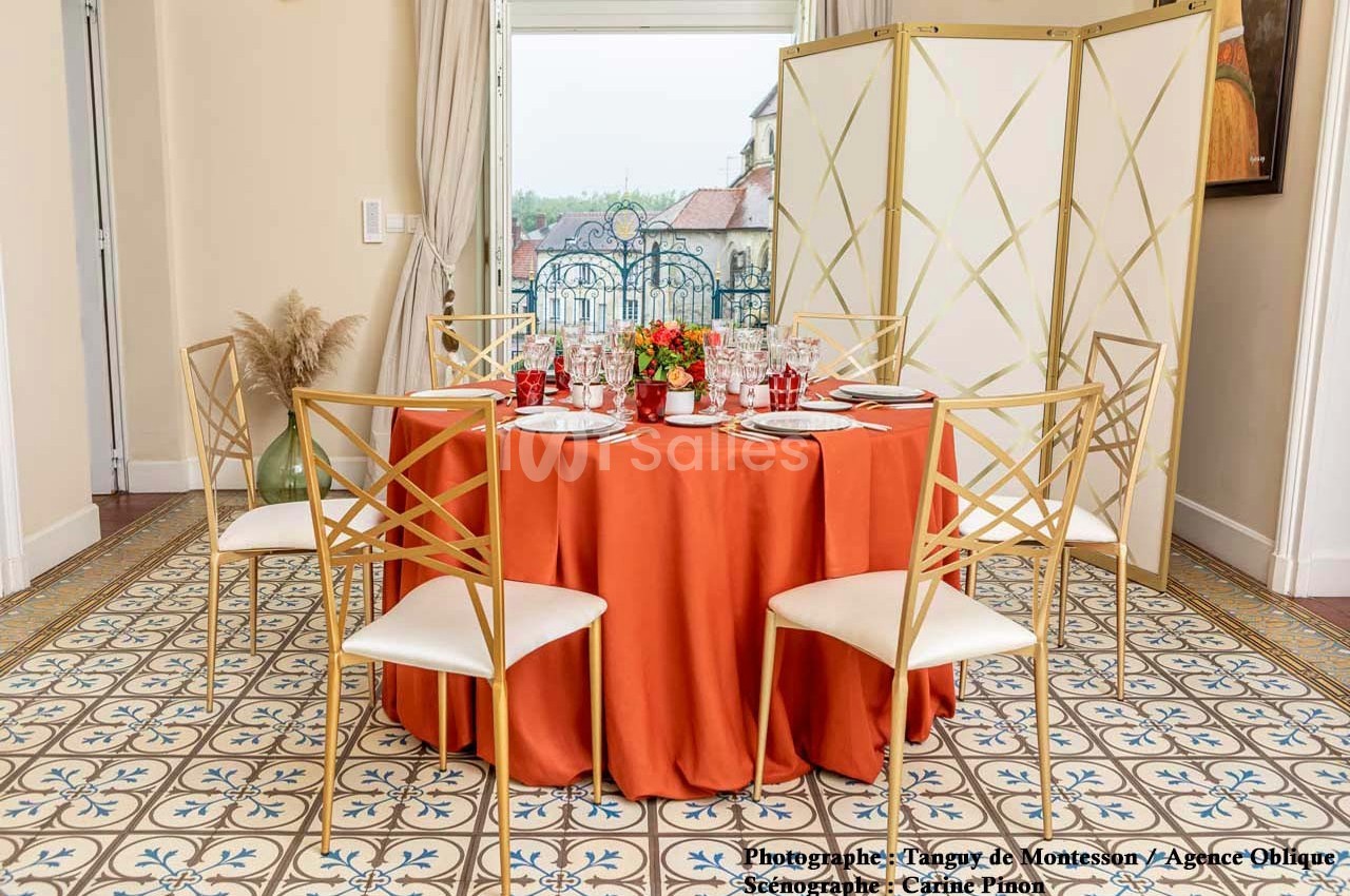 Table ronde dressée avec une nappe orange, entourée de chaises dorées, dans une pièce lumineuse au sol carrelé.