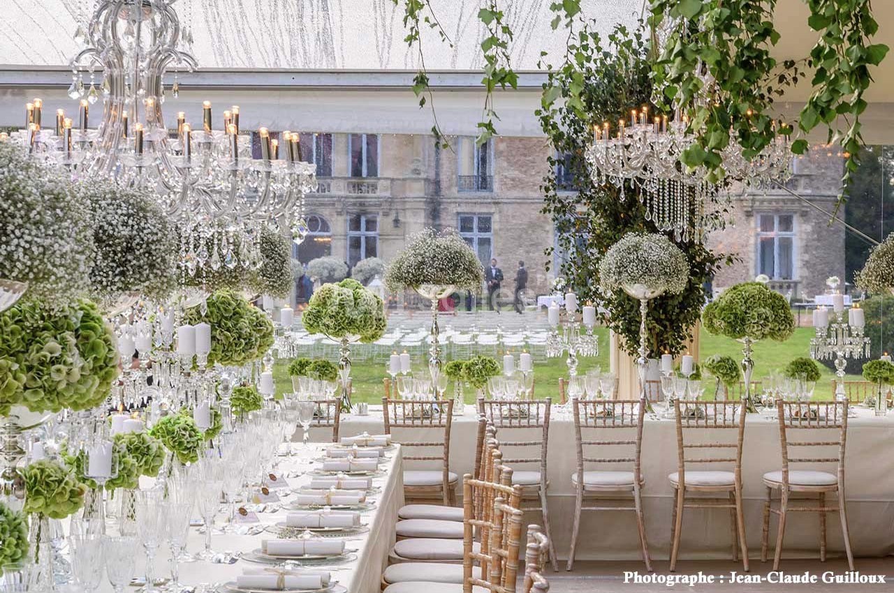 Salle de réception élégante avec tables décorées de fleurs blanches et vertes, lustres suspendus et vue sur un jardin.