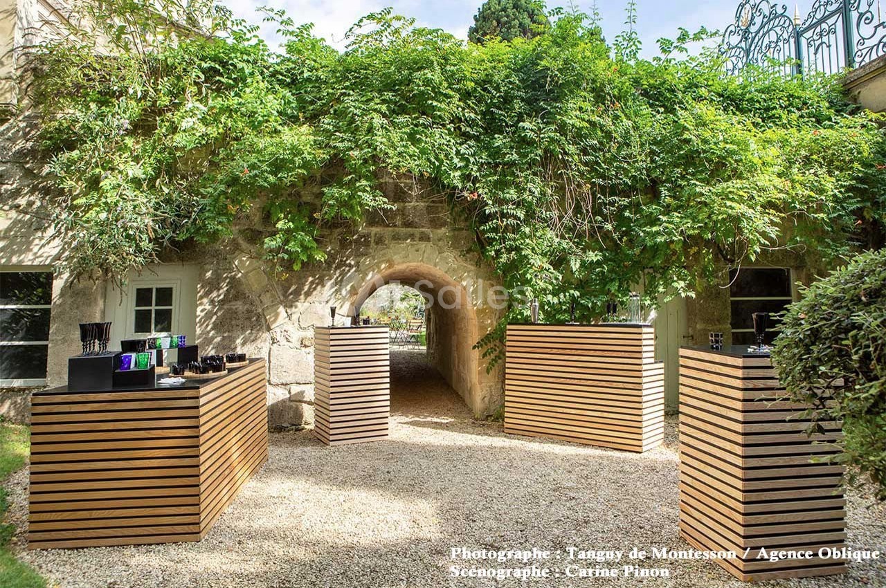 Passage voûté en pierre entouré de végétation, avec des présentoirs en bois alignés sur un chemin gravillonné.