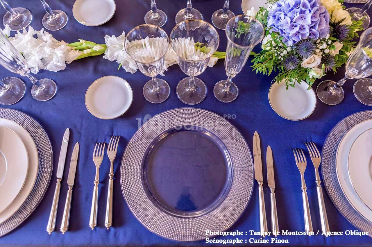 Table dressée avec vaisselle élégante, couverts en argent, verres en cristal et centre de table floral sur nappe bleue.