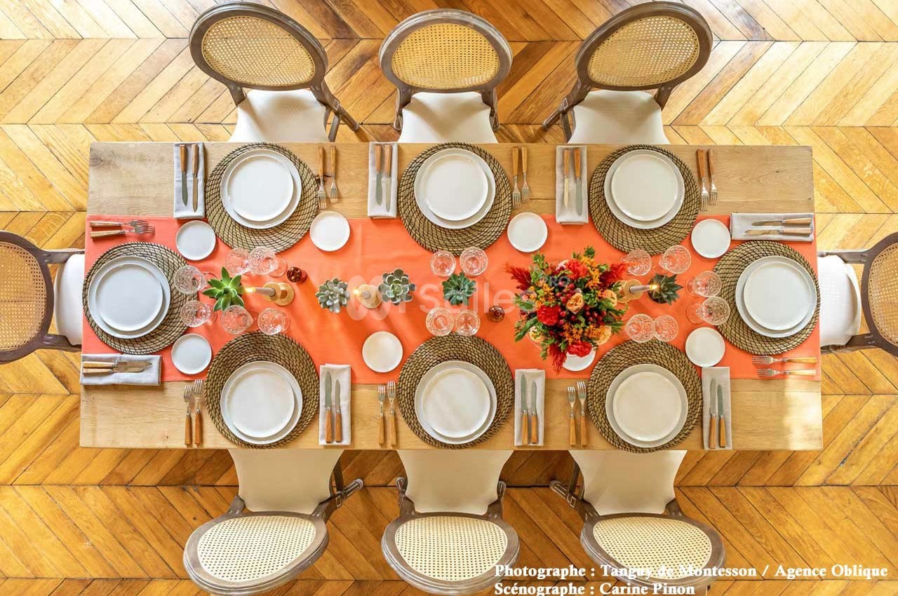 Table dressée vue de dessus avec nappes orange, assiettes blanches, sets en osier, couverts et bouquet de fleurs au centre.